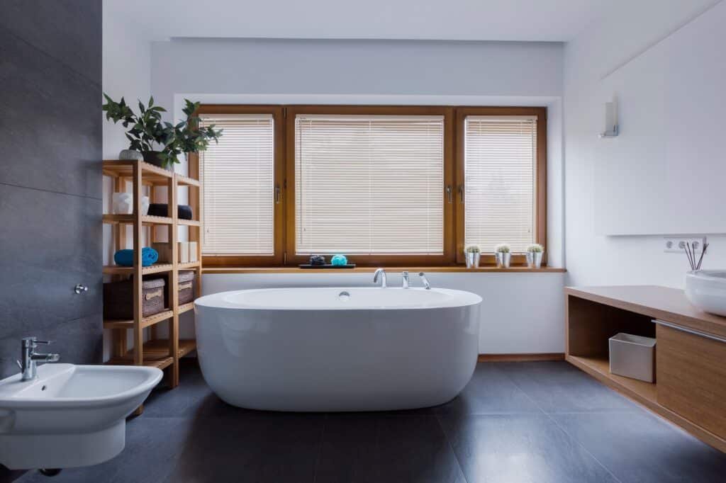 Salle de bain spacieuse à Amiens avec baignoire îlot devant fenêtre, sol carrelé foncé et meuble bas en bois.